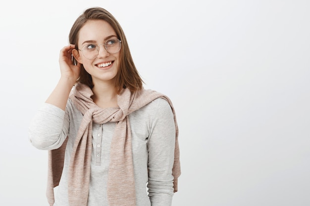 Foto da cintura para cima de uma garota feminina criativa e charmosa de óculos e pulôver tricotada no pescoço, colocando a mecha de cabelo atrás da orelha e olhando encantada e terna para a direita com um sorriso fofo