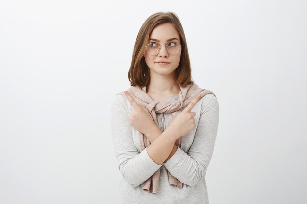 Foto da cintura para cima de uma garota feminina relaxada e bonita de óculos com cabelo castanho curto cruzando os braços no corpo apontando em lados diferentes, à esquerda e à direita, fazendo uma escolha ou decisão sobre a parede cinza