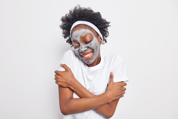 Foto da cintura para cima de uma mulher afro-americana feliz se abraçando, relembrando boas lembranças, mantém os olhos fechados, aplica máscara hidratante de argila para cuidados com a pele