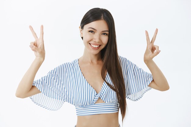 Foto da cintura para cima de uma mulher asiática feliz e amigável, com longos cabelos escuros, mostrando um gesto de paz ou vitória