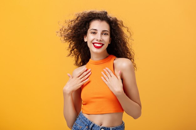 Foto da cintura para cima de uma mulher sensual e feminina tenra com penteado encaracolado em top recortado segurando as palmas das mãos no peito encantado e satisfeito sorrindo amplamente posando glamour e sexy sobre a parede laranja. Copie o espaço