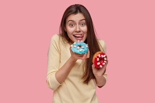 Foto da cintura para cima de uma senhora caucasiana feliz lambendo os lábios com a língua, segurando deliciosos donuts, não mantém a dieta