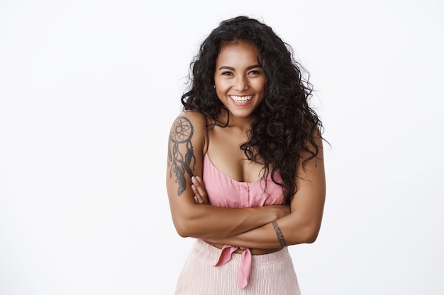 Foto da cintura para cima, feliz, despreocupada, garota atraente de cabelos encaracolados, com braços tatuados, sorriso branco cheio de dentes, peito de mãos cruzadas e câmera olhando com entusiasmo, parede branca de pé