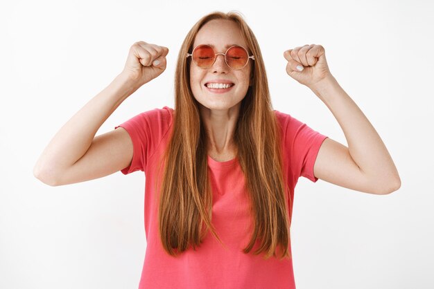 Foto da cintura para cima para triunfar feliz e emocionada jovem ruiva com sardas nos óculos escuros e camiseta rosa casual levantando os punhos cerrados na vitória, comemorando o final do semestre com sucesso