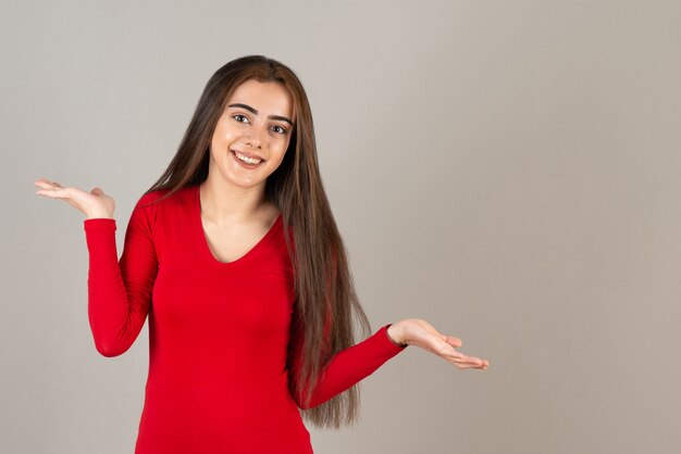 Foto da menina adorável sorridente em pé de moletom vermelho na parede cinza.