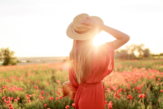 Foto da parte de trás de uma jovem inspirada segurando o chapéu de palha e olhando para o horizonte. Conceito de liberdade. Cores quentes do sol. Campo de papoula.