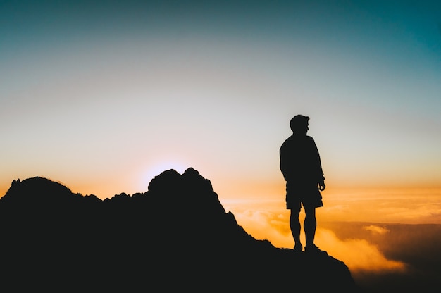 Foto da silhueta de um homem parado em um penhasco olhando o pôr do sol