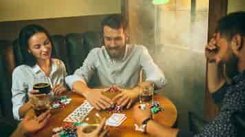 Foto grátis foto da vista lateral de amigos do sexo masculino e feminino sentados à mesa de madeira. homens e mulheres jogando jogo de cartas. mãos com close-up de álcool.