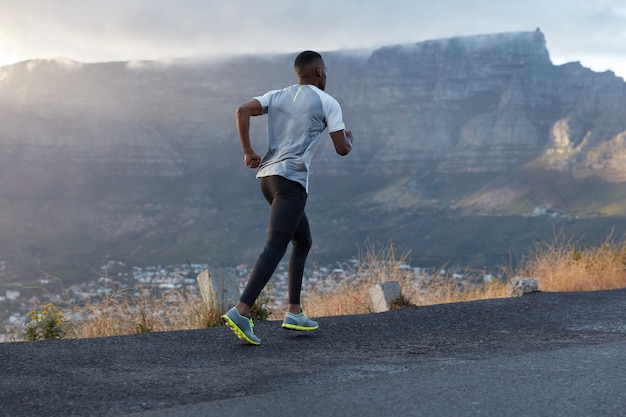 Foto das costas de um homem ativo de pele escura em ação, atravessa uma estrada de montanha, leva um estilo de vida saudável, tem resistência e motivação para estar em forma, posa sobre a montanha, gosta da natureza