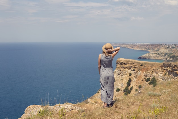 Foto grátis foto das costas de uma jovem em um vestido elegante admirando a vista deslumbrante enquanto está de pé na beira do topo da montanha, de frente para o vasto oceano azul, segurando um chapéu de palha para mantê-lo na cabeça por causa do vento