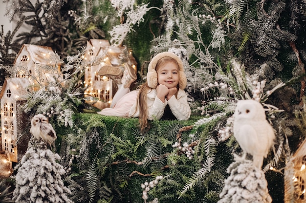 Foto de adorável criança caucasiana em protetores de ouvido bege deitado com o queixo nas mãos, rodeado por decorações de natal e sorrindo para a câmera. conceito de país das maravilhas do inverno.