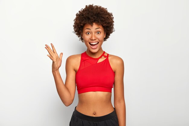 Foto de alegre mulher de pele escura levanta a palma da mão, usa sutiã esportivo vermelho, mostra a barriga, tem um penteado afro, posa interior sobre fundo branco.