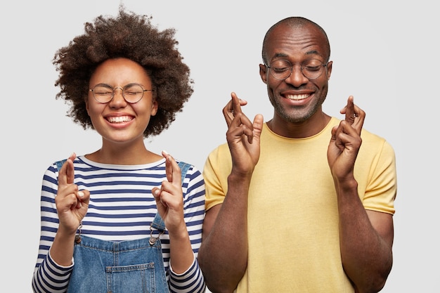 Foto de alegres esperançosos estudantes do sexo feminino e masculino de pele escura cruzando os dedos