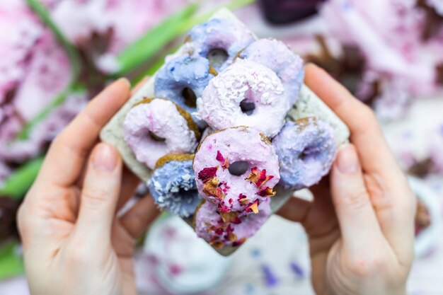 Foto de alto ângulo das mãos de uma pessoa segurando alguns donuts veganos roxos e azuis sobre uma mesa