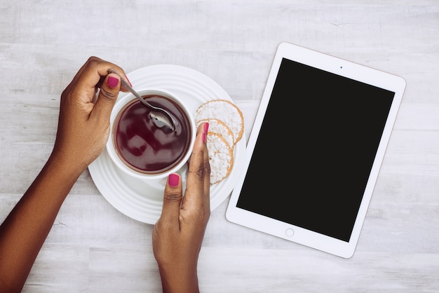 Foto de alto ângulo de uma mulher segurando uma xícara de chá com biscoitos e um tablet ao lado