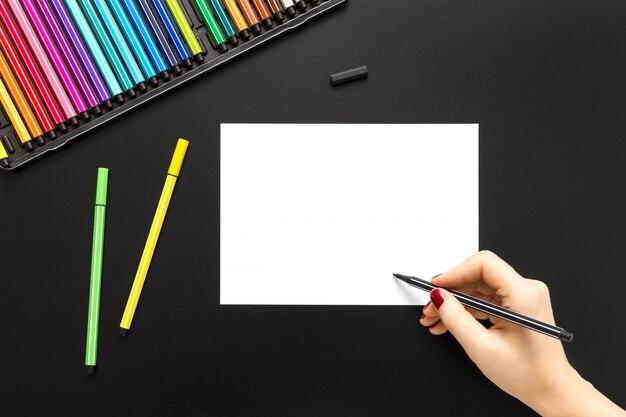 Foto de alto ângulo de uma pessoa que desenha em um papel branco com canetas coloridas em uma superfície preta