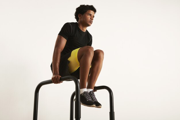 Foto de baixo ângulo de um modelo masculino preto magro e musculoso forte com um afro em roupas de treino pretas, levantando os joelhos em barras paralelas isoladas em branco.