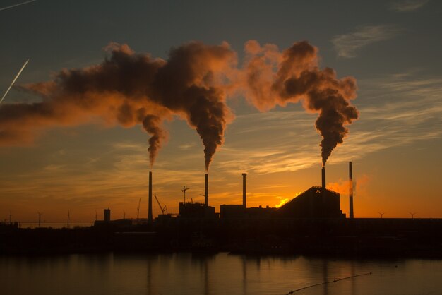 Foto de baixo ângulo de uma fábrica com fumaça saindo das chaminés capturada ao pôr do sol