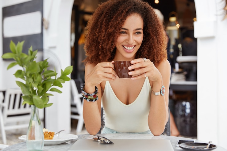 Foto grátis foto de bela sorridente mulher de pele escura com cabelo encaracolado afro bebidas expresso no café tem uma expressão positiva.
