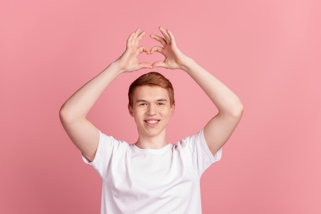Foto de cara jovem demonstra dedos em forma de coração, amor romântico, isolado sobre fundo de cor rosa