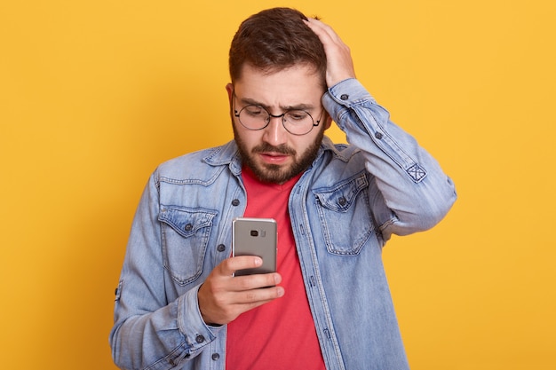 Foto de carismático chocado barbudo jovem segurando seu smartphone, olhando atentamente a tela do dispositivo, colocando a mão na cabeça