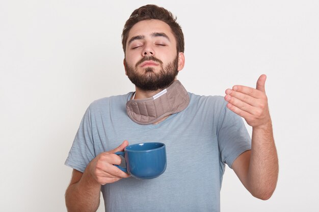 Foto de carismático homem jovem e bonito, fechando os olhos, segurando o copo com uma bebida quente, apreciando o cheiro de café, vestindo pijama