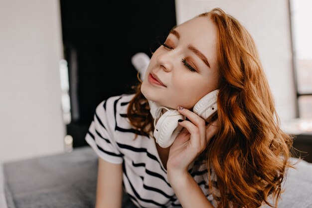 Foto de close-up de alegre mulher de gengibre com maquiagem da moda. Garota Jocund em t-shirt listrada, posando com fones de ouvido.