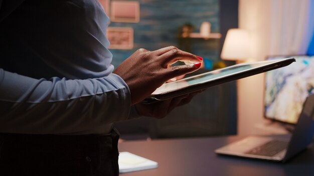 Foto de close-up de mãos femininas negras segurando um computador tablet em pé na sala de estar tarde da noite. Mulher afro-americana usando rede social, mensagens de texto e blogs fazendo hora extra por trabalho
