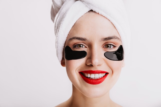 Foto de close-up de menina de olhos verdes com adesivos hidratantes. Jovem mulher com lábios vermelhos, sorrindo sobre uma parede branca.