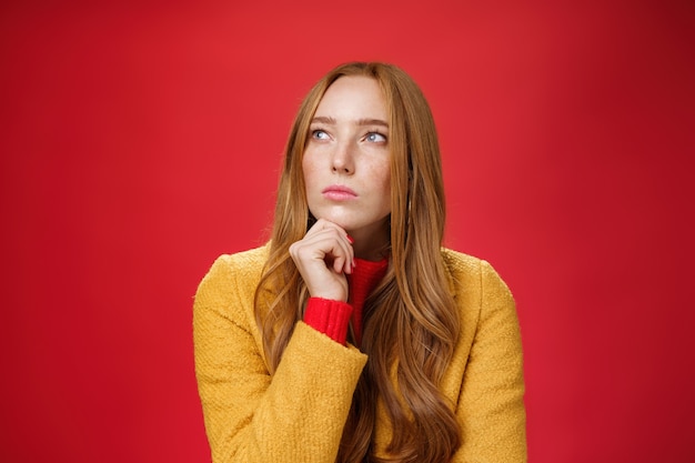 Foto de close-up de mulher ruiva pensativa criativa determinada e focada olhando para o canto superior esquerdo tocando o queixo, pensando, fazendo escolhas ou lembrando informações sobre fundo vermelho.