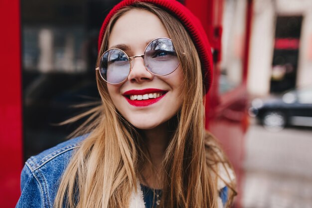 Foto de close-up de romântica garota branca de óculos redondos, olhando para cima com um sorriso. Uma jovem sonhadora com maquiagem brilhante, posando ao lado do ônibus vermelho.