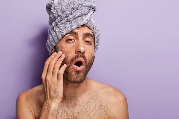 Foto de close-up de um homem bonito com a barba por fazer com máscaras sob os olhos, olha para a câmera com a boca bem aberta, corpo nu, toalha enrolada na cabeça