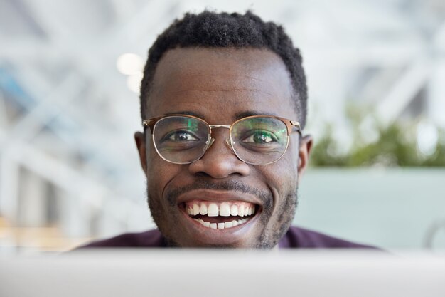 Foto de close-up de um homem feliz de pele escura com sorriso largo, dentes brancos, usando óculos transparentes para uma boa visão