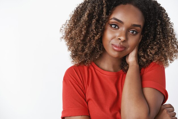 Foto de close-up de uma amiga afro-americana romântica e gentil, usando uma camiseta casual vermelha, tocando a nuca tímida e linda cabeça inclinando-se, sorrindo sensualmente com olhar sedutor sobre a parede branca