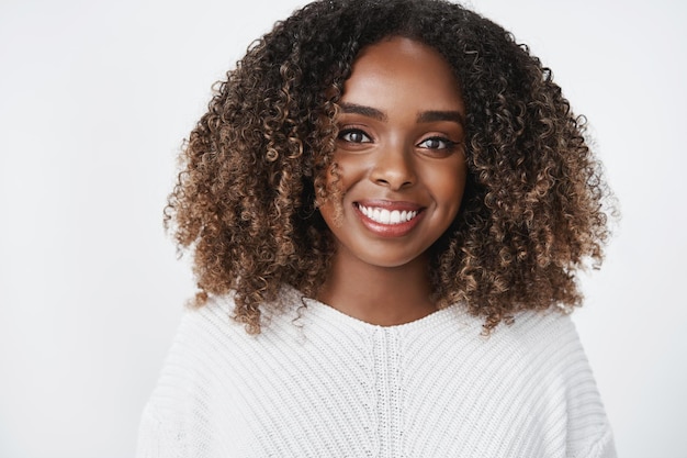 Foto de close-up de uma jovem aluna afro-americana relaxada e alegre, usando um suéter e um corte de cabelo encaracolado, sorrindo maravilhada Foto gratuita