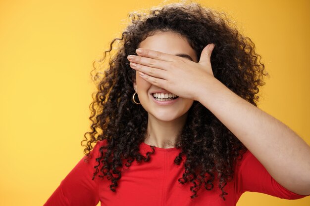 Foto de close-up de uma jovem fofa feliz sonhadora com cabelos cacheados cobrindo os olhos com a palma da mão enquanto contando ou jogando esconde-esconde sorrindo amplamente antecipando a surpresa acontecer sobre fundo amarelo.