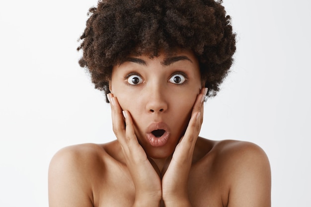 Foto de close-up de uma linda mulher afro-americana nua, animada e espantada, com cabelo encaracolado, perdendo a fala em notícias surpreendentes, segurando as mãos nas bochechas, dobrando os lábios e olhando impressionado