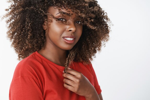 Foto de close-up de uma linda mulher de pele escura romântica e sensual com pele pura e limpa, sorrindo carinhosamente, tocando cachos de cabelo e acenando com um corte de cabelo olhando suave e carinhoso na frente sobre a parede branca