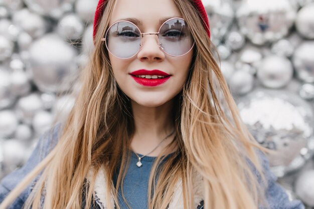 Foto de close-up de uma menina branca de óculos, expressando interesse. Senhora alegre com lábios vermelhos, posando na parede do brilho.