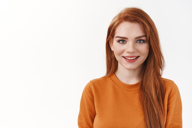 Foto de close-up, mulher ruiva sorridente, muito carismática, com suéter laranja, olhar para a frente confiável com um sorriso fofo, parede branca de pé alegre, promover um bom serviço confiável