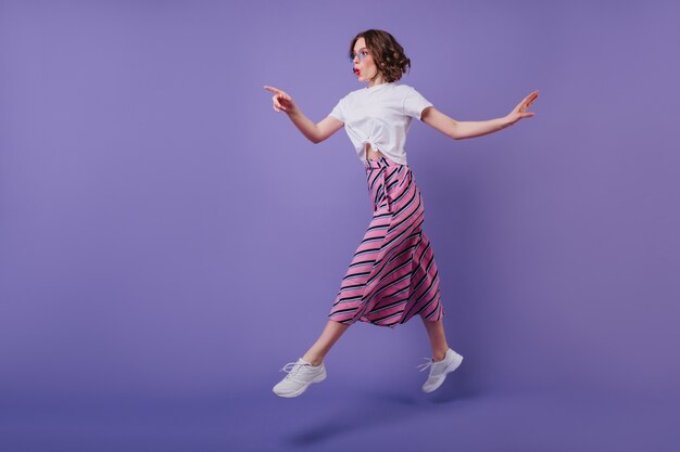 Foto de corpo inteiro de animada menina bonita com penteado ondulado, pulando na parede roxa. Mulher elegante e bem humorada de tênis se divertindo durante a sessão de fotos.