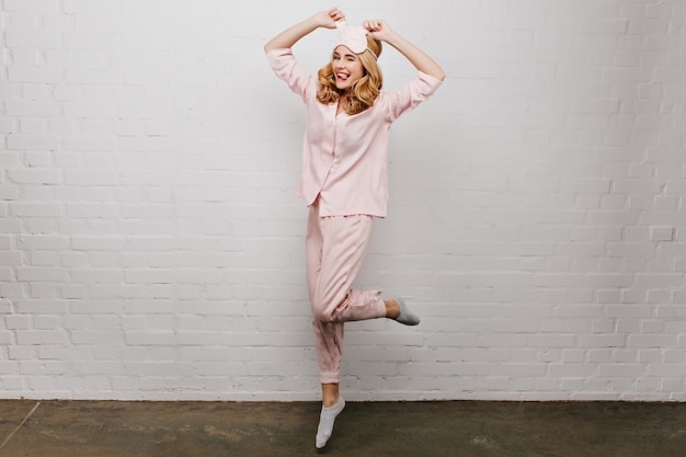 Foto de corpo inteiro de graciosa garota despreocupada, aproveitando a manhã. A deslumbrante modelo feminina usa meias cinza e pijama rosa dançando em casa.