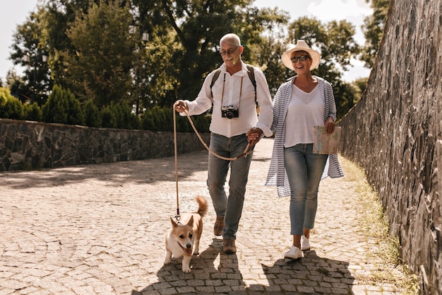 Foto de corpo inteiro de homem de camisa branca e calça jeans com câmera e mulher de chapéu e blusa azul listrada com cartão e corgi caminhando no parque.