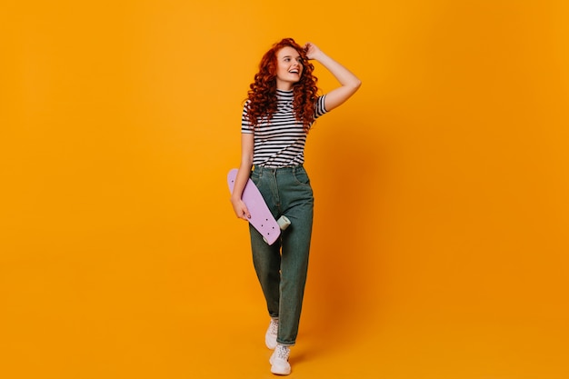 Foto de corpo inteiro de menina adolescente vermelho encaracolado com longboard nas mãos. Mulher em calças jeans e camisa posando no espaço laranja.