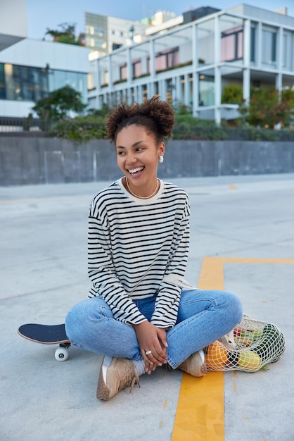 Foto de corpo inteiro de uma adolescente relaxada e feliz descansando depois que o esporte ativo está de bom humor e gosta de andar de skate, senta-se com as pernas cruzadas na estrada em um ambiente urbano e olha para longe positivamente