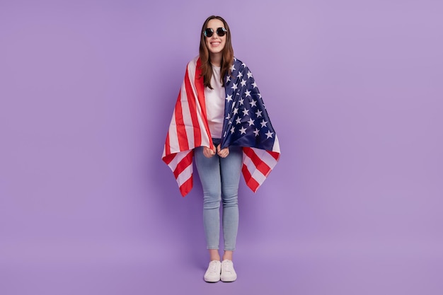 Foto de corpo inteiro de uma jovem bonita com sorriso positivo e feliz, se cobrindo com a nacionalidade da bandeira dos eua isolada sobre o fundo violeta