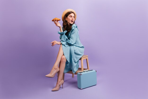 Foto de corpo inteiro de uma jovem elegante vestido azul. Mulher se senta no banquinho ao lado da mala e segura um delicioso croissant.