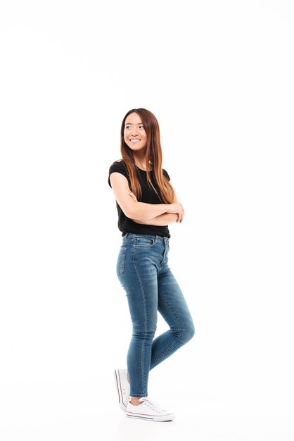 Foto de corpo inteiro de uma jovem mulher bonita chinesa em camiseta preta e calça jeans em pé com as mãos cruzadas, olhando de lado