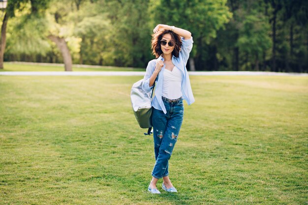 Foto de corpo inteiro de uma linda garota morena com cabelo curto em óculos de sol, posando no parque. Ela usa camiseta branca, camisa azul e jeans, sapatos, bolsa. Ela está sorrindo para a câmera.