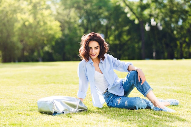 Foto de corpo inteiro de uma linda garota morena com cabelo curto, posando na grama na luz do sol no parque. Ela usa camiseta branca, camiseta e jeans, sapatos, bolsa. Ela está sorrindo para a câmera.